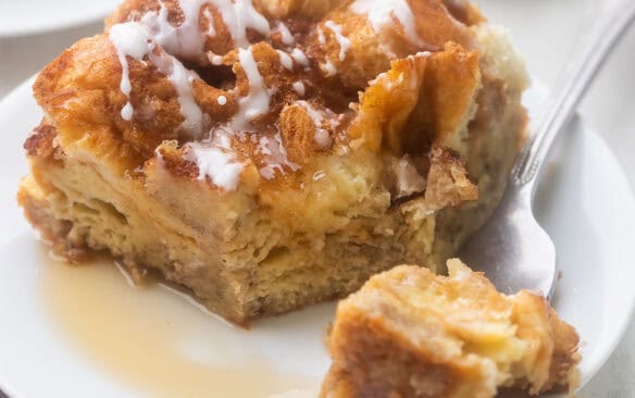 close up of a piece of french toast casserole on a white plate with a forkful beside.