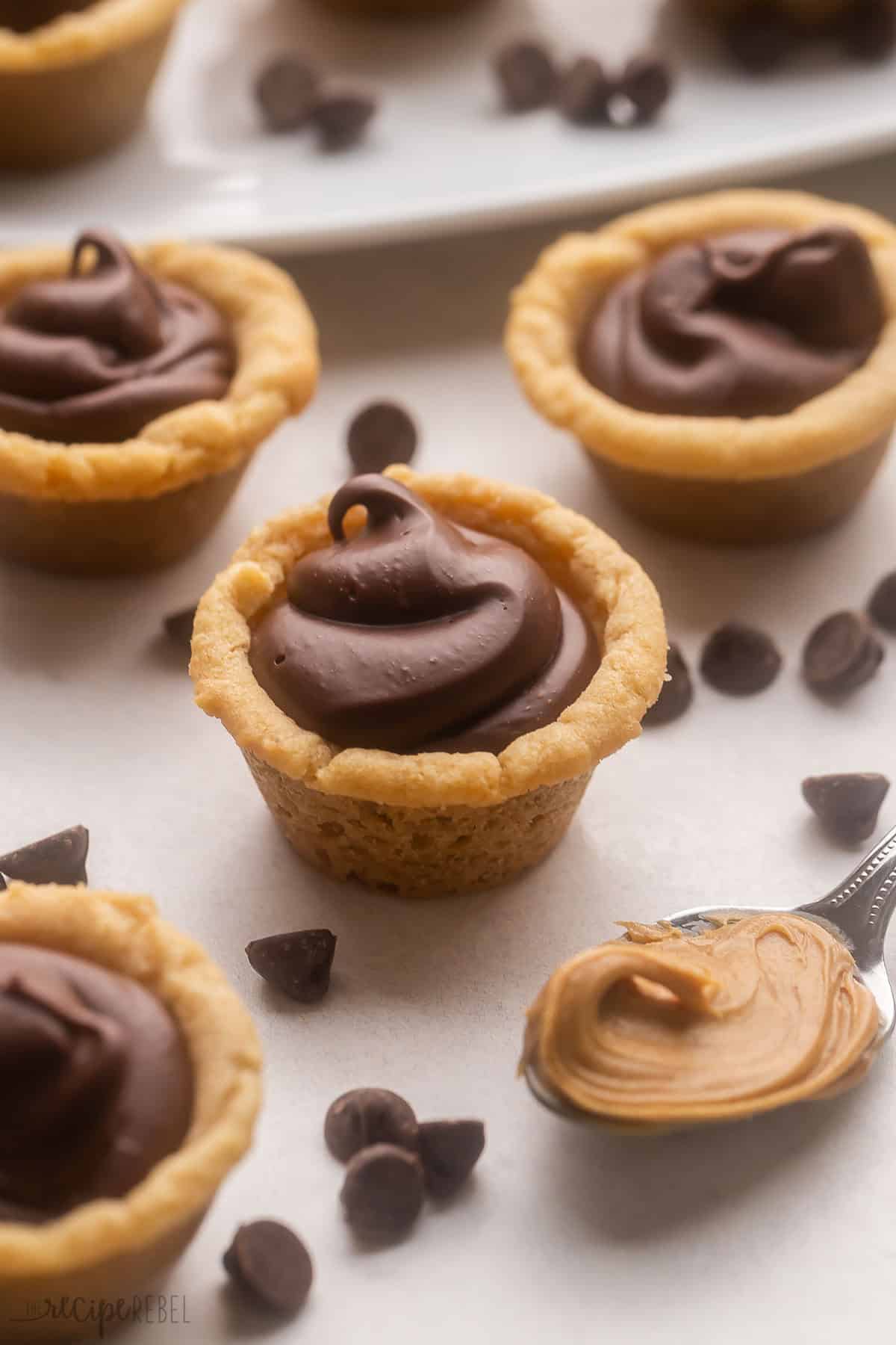 Chocolate Peanut Butter Cookie Cups, or more affectionately known as Fudge Puddles, are made with a peanut butter cookie dough base and a fudgy center made of chocolate chips and condensed milk. It's an easy dessert for curbing those peanut butter chocolate cravings or for enjoying during the holiday season! #chocolate #peanutbutter #cookies | christmas cookies | christmas treat | holiday baking | peanut butter cups | cookie cup | easy dessert | dessert recipe fudge puddles and a spoonful of peanut butter surrounded by chocolate chips.