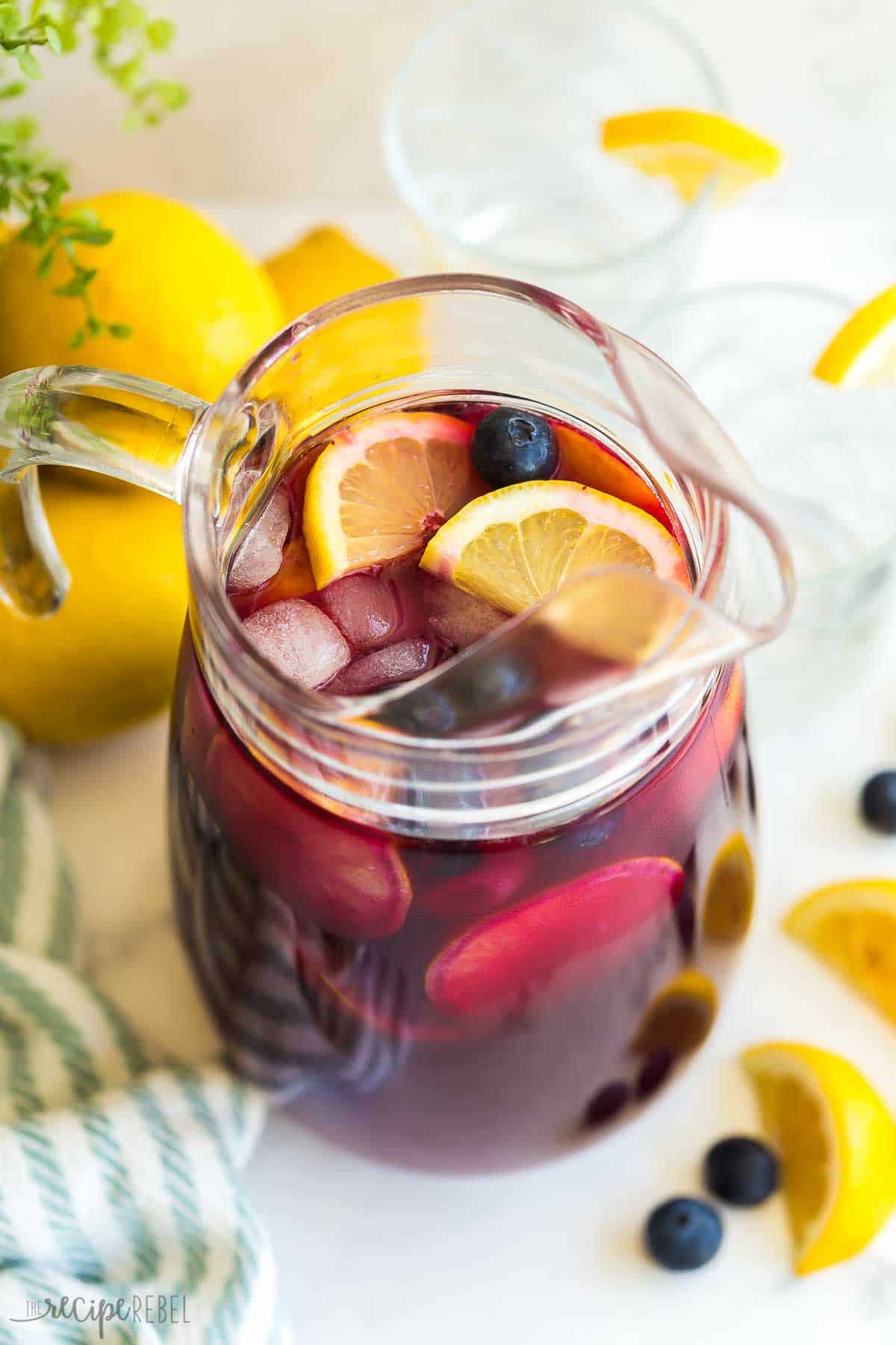 This Blueberry Lemonade is sweet, tangy, and perfect for summer. This refreshing drink is made with fresh lemon and blueberry lemon simple syrup! #lemon #summer #drinks | lemonade recipe | homemade lemonade | simple syrup | lemon recipes | blueberry recipes | blueberries | drinks glass pitcher of blueberry lemonade with lemons in the background.