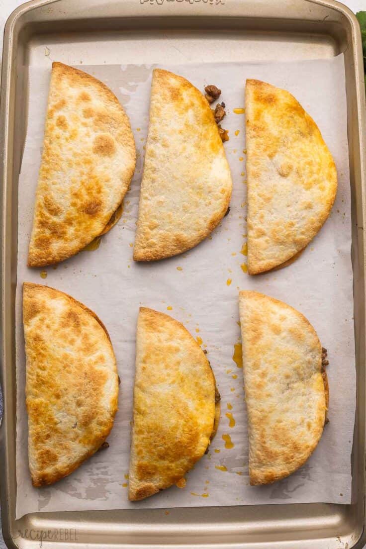 six baked tacos lying in a parchment lined pan.