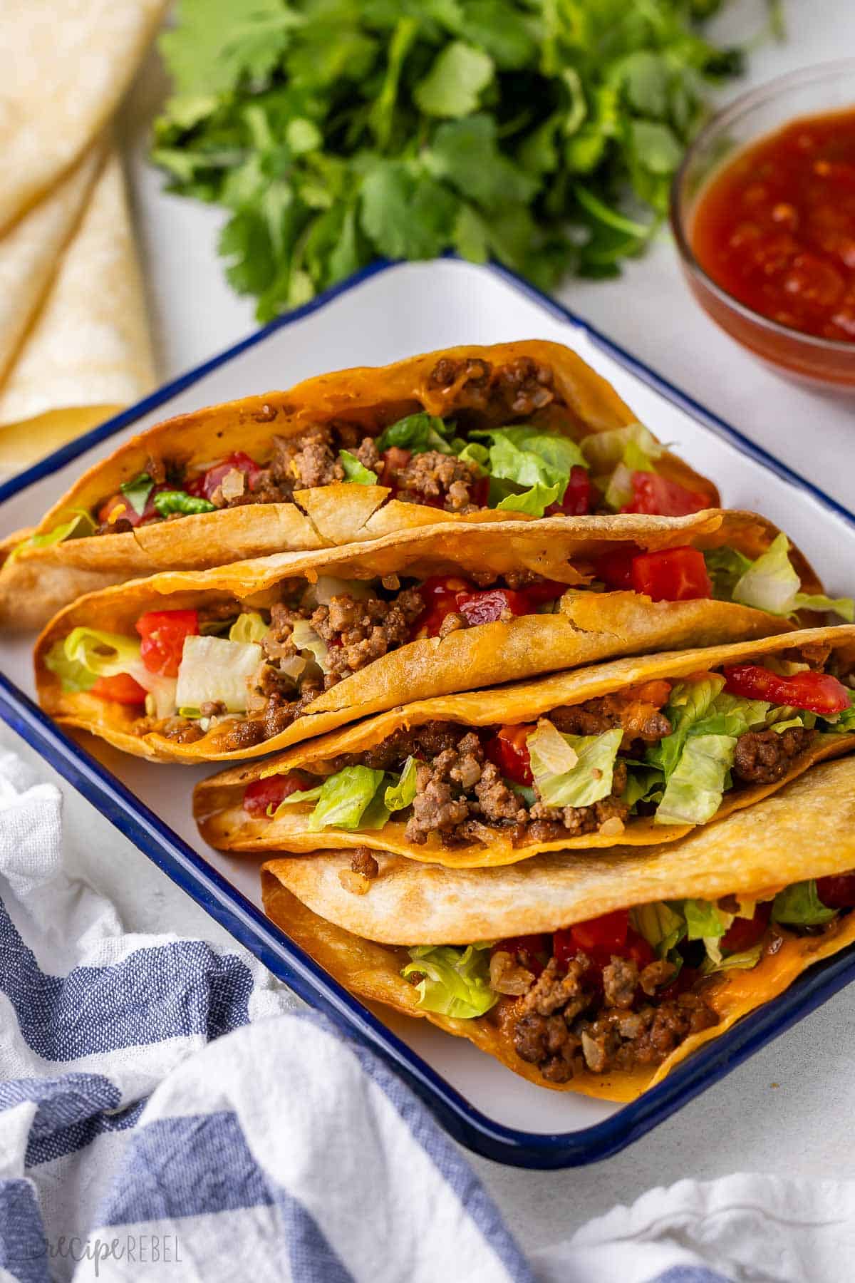 These Crispy Baked Tacos are filled with a juicy ground beef mixture and melted cheese and baked until golden brown. They're made with simple ingredients and ready in 40 minutes, perfect for game day or as an easy dinner! #dinner #recipe #taco | ground beef recipes | ground beef dinner | ground beef tacos | baked tacos | taco recipes | easy dinner | dinner ideas | quick meals crispy baked tacos with parsley and salsa behind it.