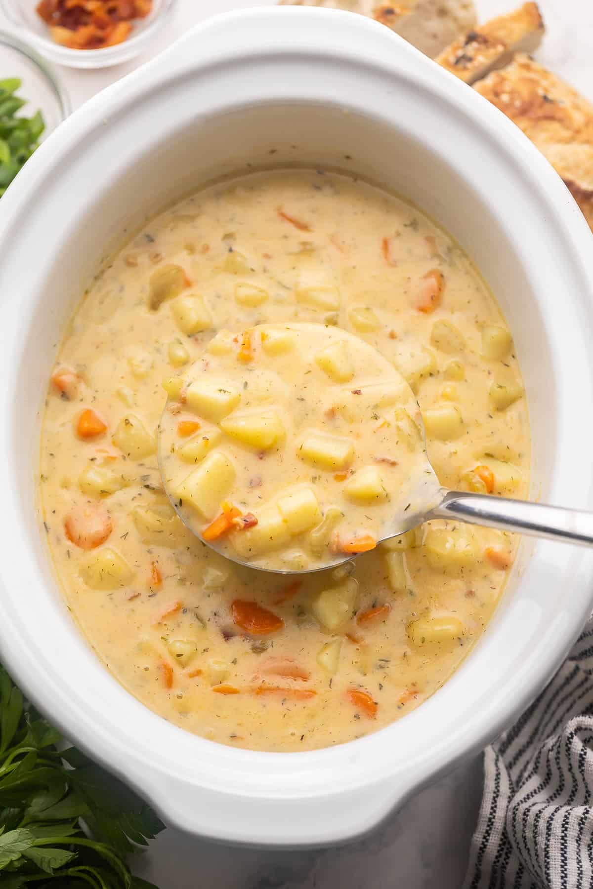 This Crockpot Potato Soup is thick, creamy, and loaded with tender potatoes, crispy bacon, cheddar cheese, green onions, and a few veggies for good measure. Cooked in the slow cooker for hours, it’s a great recipe for busy days! #slowcooker #crockpot #soup #recipe | crockpot meals | slow cooker soup | potato recipes | comfort food | soup recipes ladle scooping potato soup out of crockpot.