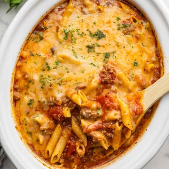 white slow cooker filled with baked ziti and wooden ladle.
