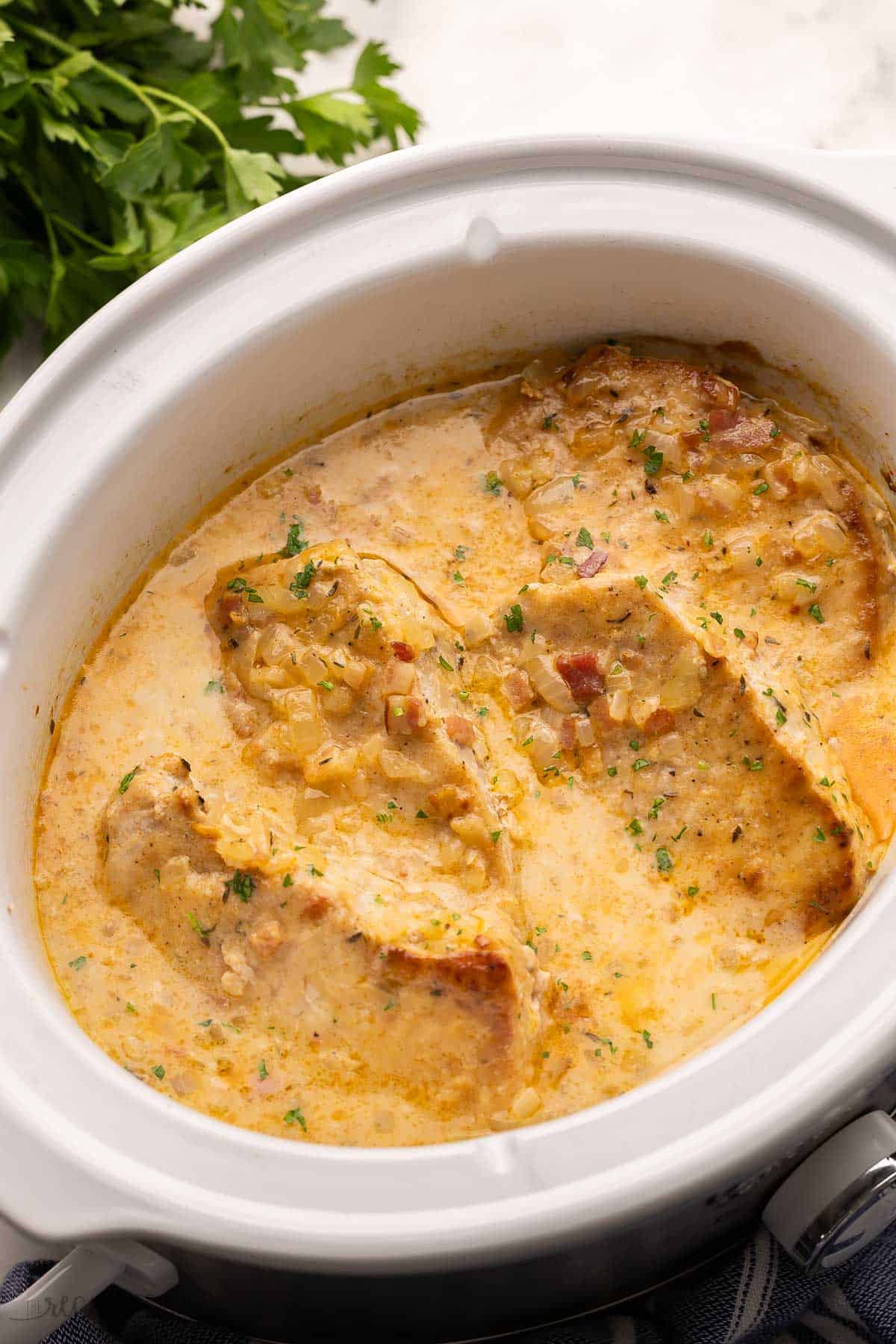 pork chops and sauce in white crockpot with parsley behind it.