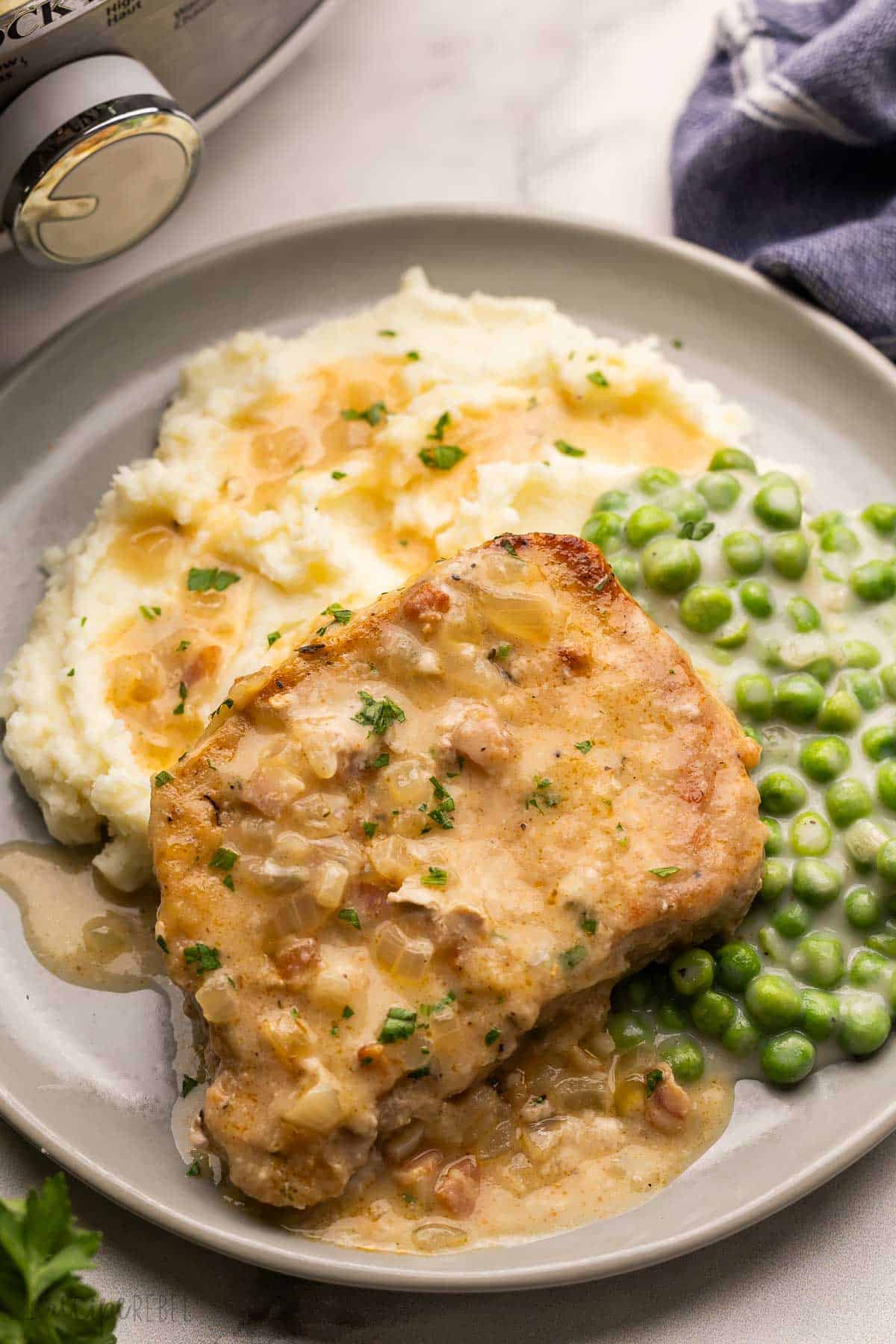 Slow Cooker Smothered Pork Chops are a Southern-inspired meal made with juicy thick cut pork chops simmered in a rich, flavorful gravy in the crockpot. It's the perfect comfort food meal for busy days when you need a hands-off meal! #slowcooker #crockpot #dinner | dinner ideas | dinner recipes | slow cooker meals | crockpot meals | crock pot recipes | boneless pork chops | pork chops recipe | crockpot pork chops | comfort food | gravy a gray plate filled with mashed potatoes, peas and a pork chop.