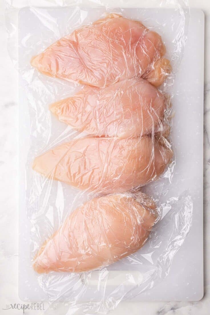 four chicken breast on a white cutting board covered in plastic wrap.