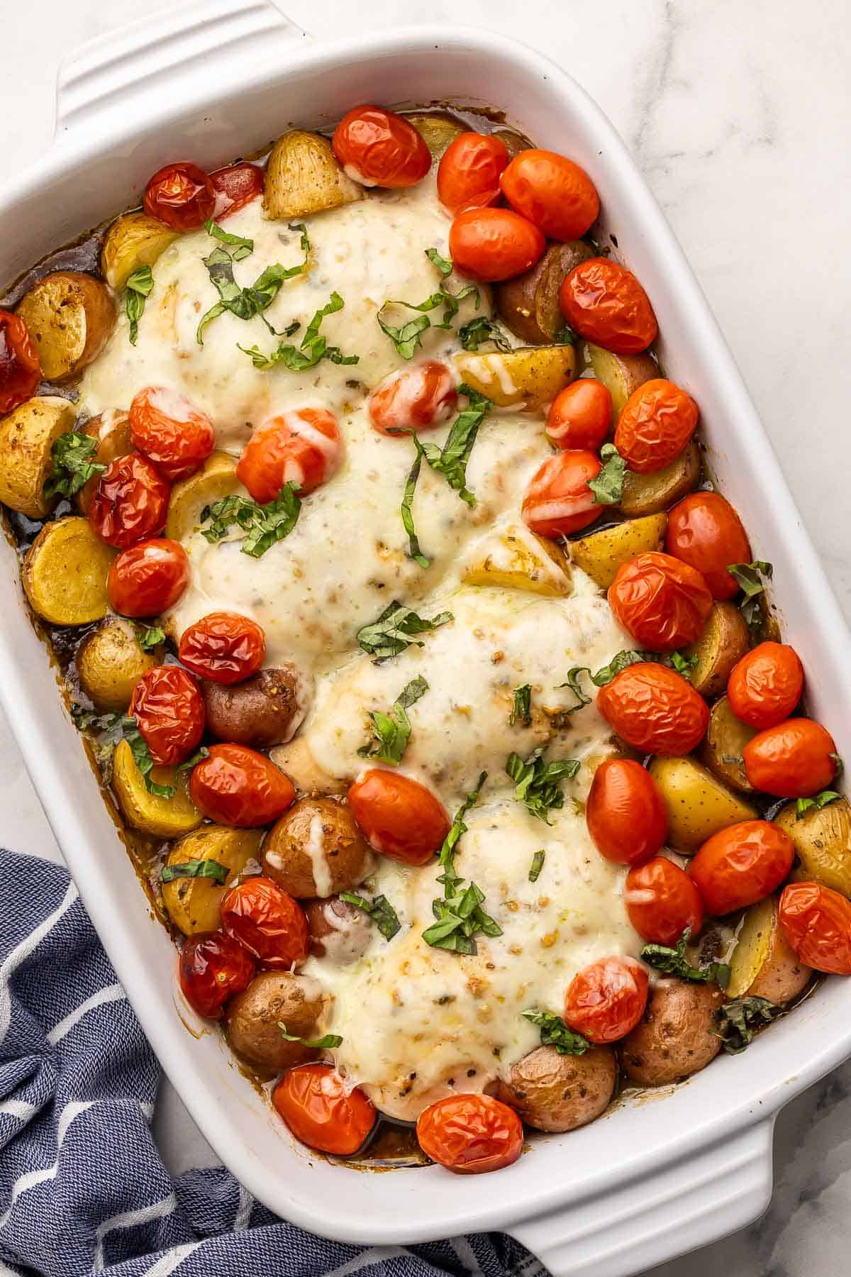 This Caprese Chicken and Potatoes is a simple casserole packed with big flavor! Chicken, potatoes, and tomatoes bake with Italian herbs and balsamic, then get topped with mozzarella and fresh basil for an easy, cozy meal. #dinner #recipe #chicken | chicken dinner | chicken recipes | balsamic | tomatoes | casserole recipe | baked chicken breast | chicken breast ideas large pan of baked caprese chicken and potatoes with chopped basil on top.