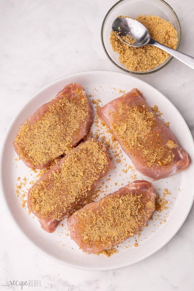 four pork chops on a plate covered with rub ingredients.
