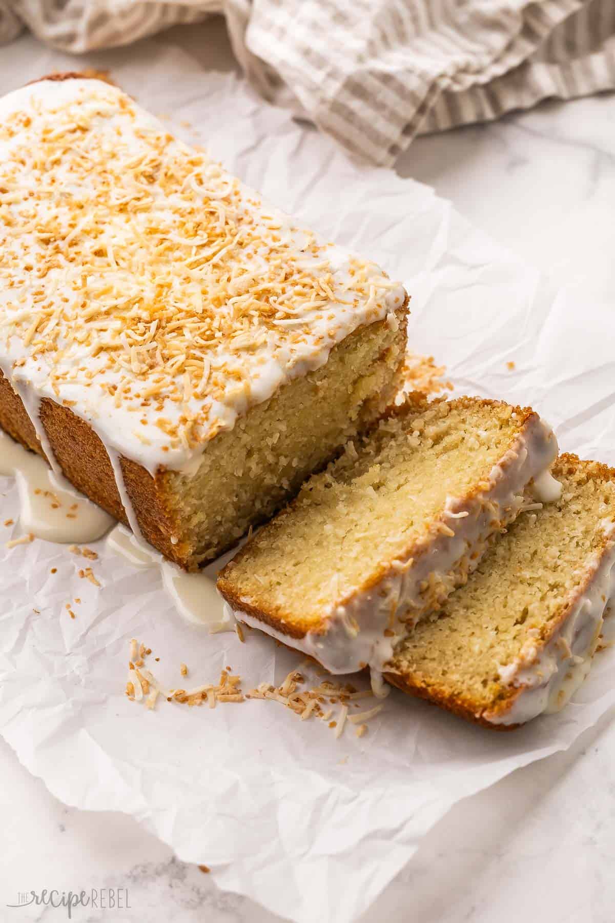 This moist Coconut Bread is made with coconut milk, coconut extract, shredded coconut, Greek yogurt, and pantry staples for a quick tropical treat! It's topped with a rich cream cheese glaze for the perfect finishing touch. #easter #baking #bread | baking recipes | easter brunch | easter dessert | easter recipes | easter treats | coconut recipe | quick bread | spring desserts coconut bread on parchment paper with a couple of slices cut.