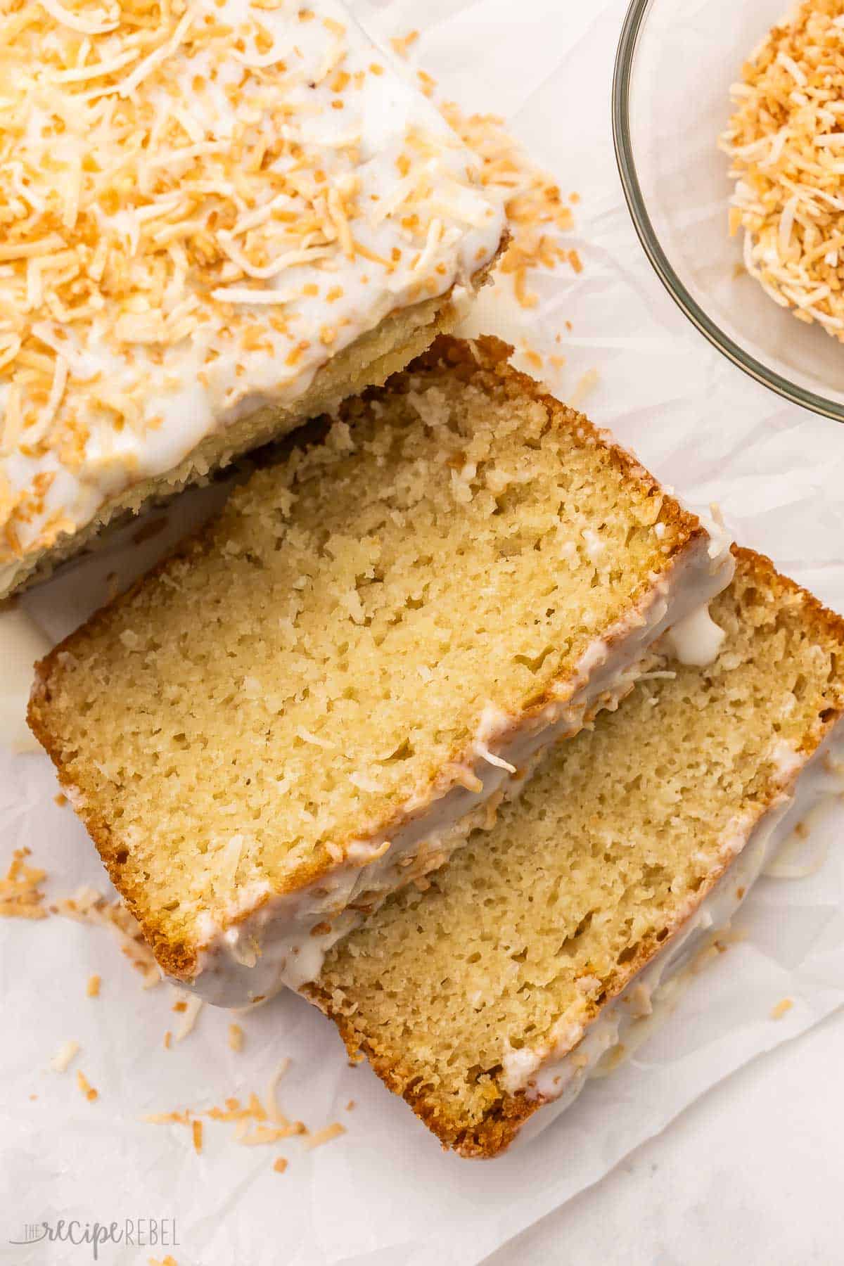 This moist Coconut Bread is made with coconut milk, coconut extract, shredded coconut, Greek yogurt, and pantry staples for a quick tropical treat! It's topped with a rich cream cheese glaze for the perfect finishing touch. #easter #baking #bread | baking recipes | easter brunch | easter dessert | easter recipes | easter treats | coconut recipe | quick bread | spring desserts two slices of coconut bread on white parchment paper.