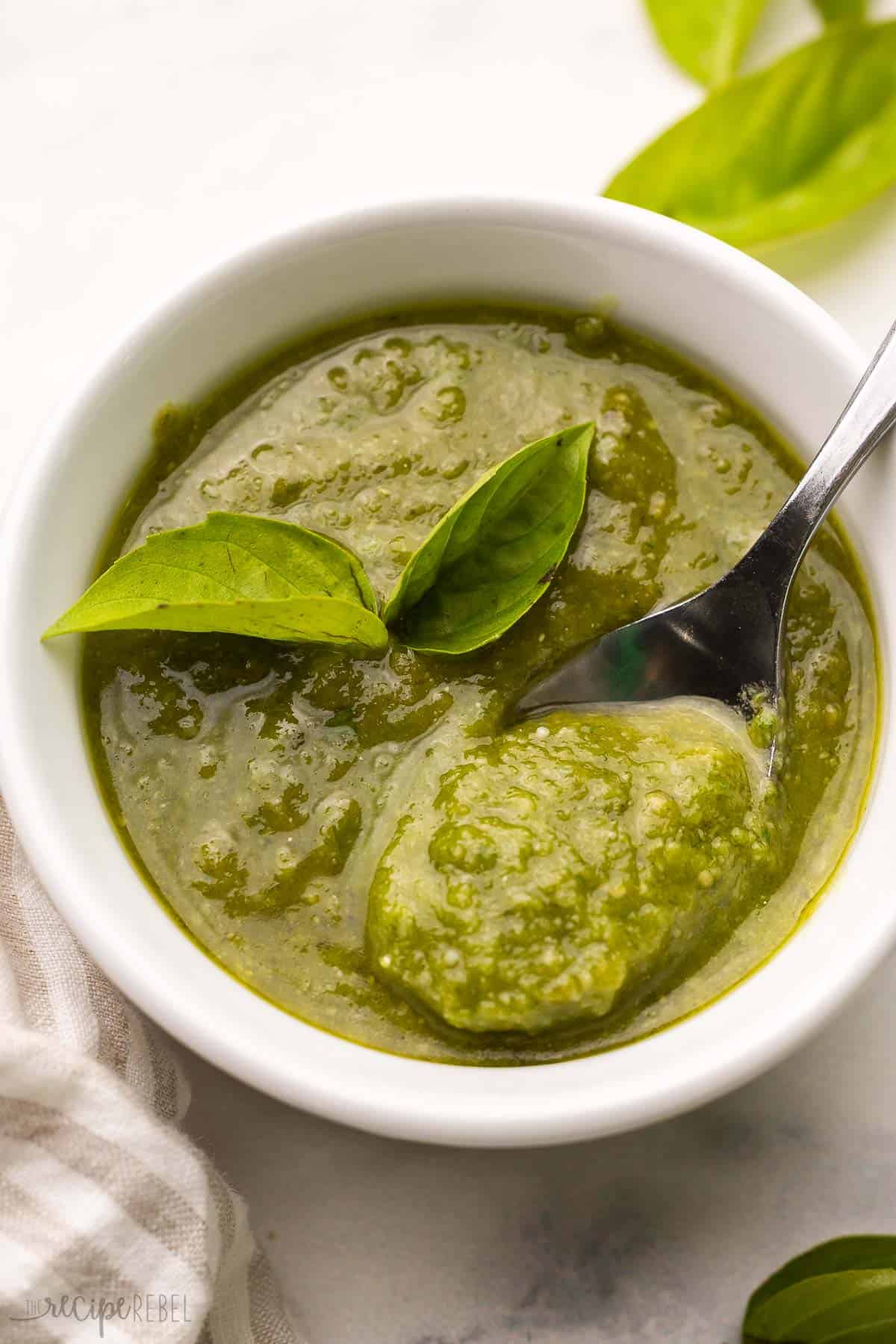 Elevate any dish with this homemade Basil Pesto recipe! It's loaded with a ton of fresh basil, olive oil, parmesan cheese, pine nuts, and garlic for a punch of fresh flavor. Perfect for topping pasta, bread, salad, or anything else you like. #recipe | pesto recipe | fresh basil | basil recipes | homemade pesto recipe | fresh herbs white bowl full of basil pesto with a spoon in it.