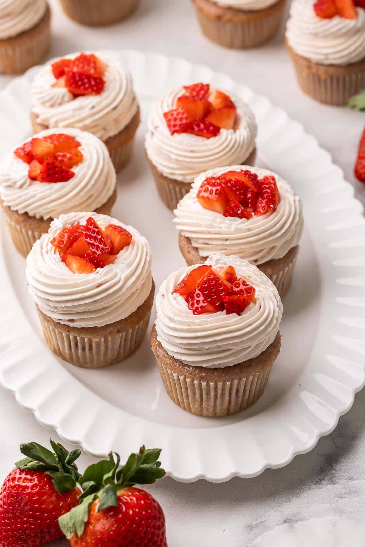 These Strawberry Cupcakes are made with real strawberries and topped with a creamy strawberry whipped cream. They're perfect for any summer birthday, party, or baby shower with their pretty pink hue! #cupcakes #dessert #baking | strawberry dessert | baking recipes | cupcake recipes | summer dessert | strawberry dessert six strawberry cupcakes on a white platter with strawberries beside.