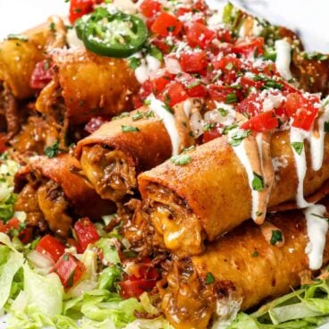 beef and cheese taquitos piled on top of each other on top of a bed of lettuce topped with sour cream, jalapeños, and pico de Gallo.