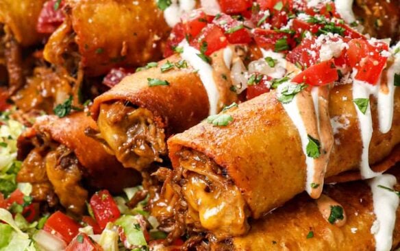 beef and cheese taquitos piled on top of each other on top of a bed of lettuce topped with sour cream, jalapeños, and pico de Gallo.