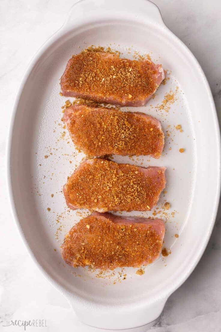 four pork chops in white baking dish with seasoning rub on them.
