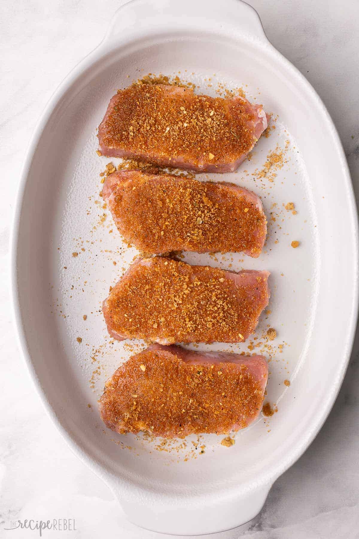four pork chops in white baking dish with seasoning rub on them.