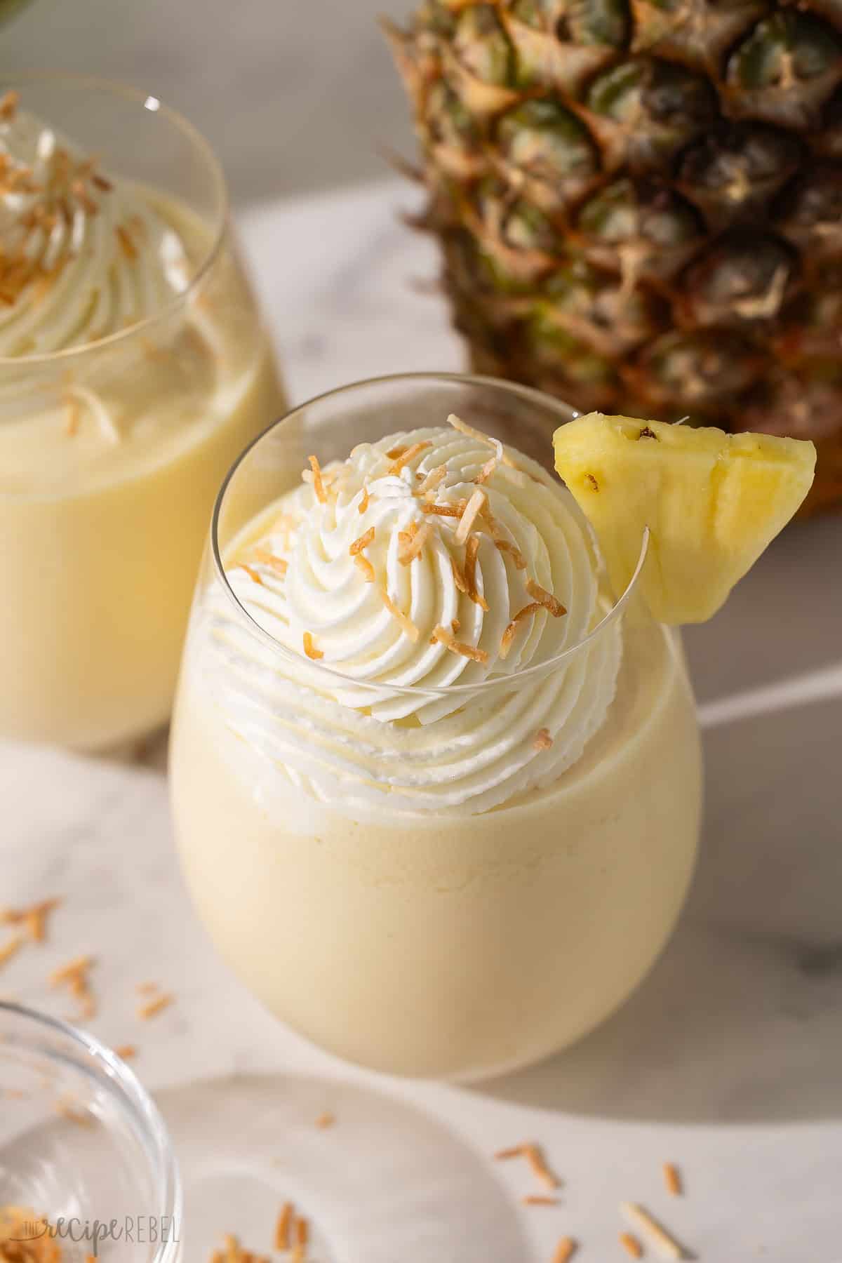 Get a taste of the tropics with this Frozen Pina Colada slushy recipe! It's made with creamy coconut milk, condensed milk, vanilla, frozen pineapple, and tons of ice for a drink that feels like a beach vacation. #drink #recipe #mocktail | mocktail recipe | drink recipe | summer drink | frozen drinks | blended drink recipe glass of pina colada slushy with a pineapple behind it.