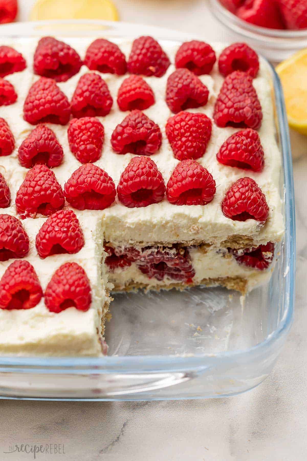 Lemon Raspberry Icebox Cake layers crisp graham crackers, lemon curd whipped cream, and heaps of fresh raspberries for an effortless no-bake treat. Perfect for warm spring and summer days! #lemon #dessert #recipe | lemon recipes | lemon dessert recipe | easy dessert | no bake dessert | raspberry recipes | summer dessert recipe a pan of lemon raspberry icebox cake with a piece missing.
