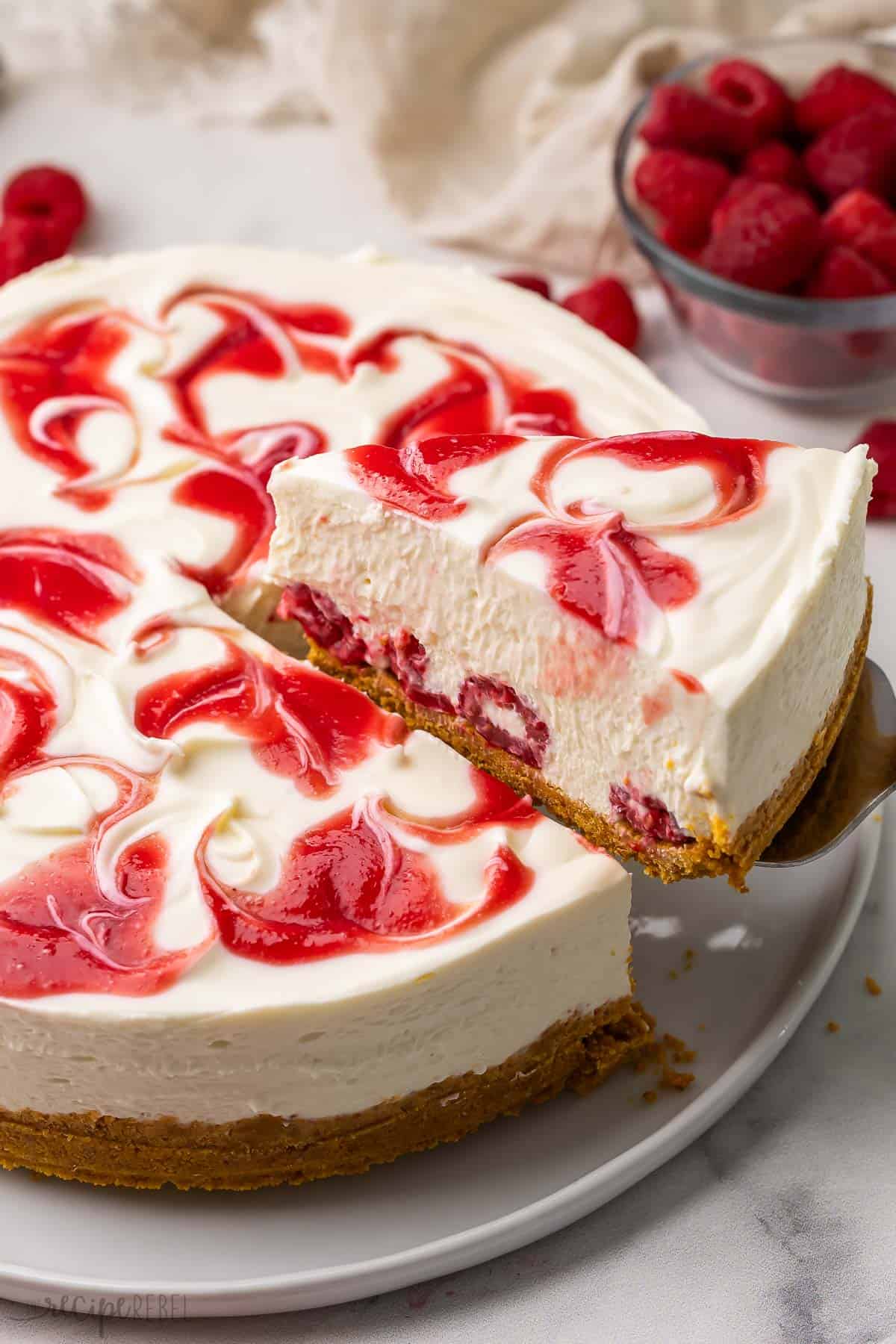This No Bake White Chocolate Raspberry Cheesecake has a creamy white chocolate cheesecake filling and crunchy graham cracker crust and is swirled with a homemade raspberry sauce. Perfect for parties, potlucks, or just because! #nobake #dessert #recipe | no bake cheesecake | no bake dessert | no bake summer dessert | summer recipes | raspberry recipes | cheesecake recipes | easy dessert a piece of cheesecake being lifted out of the cake.