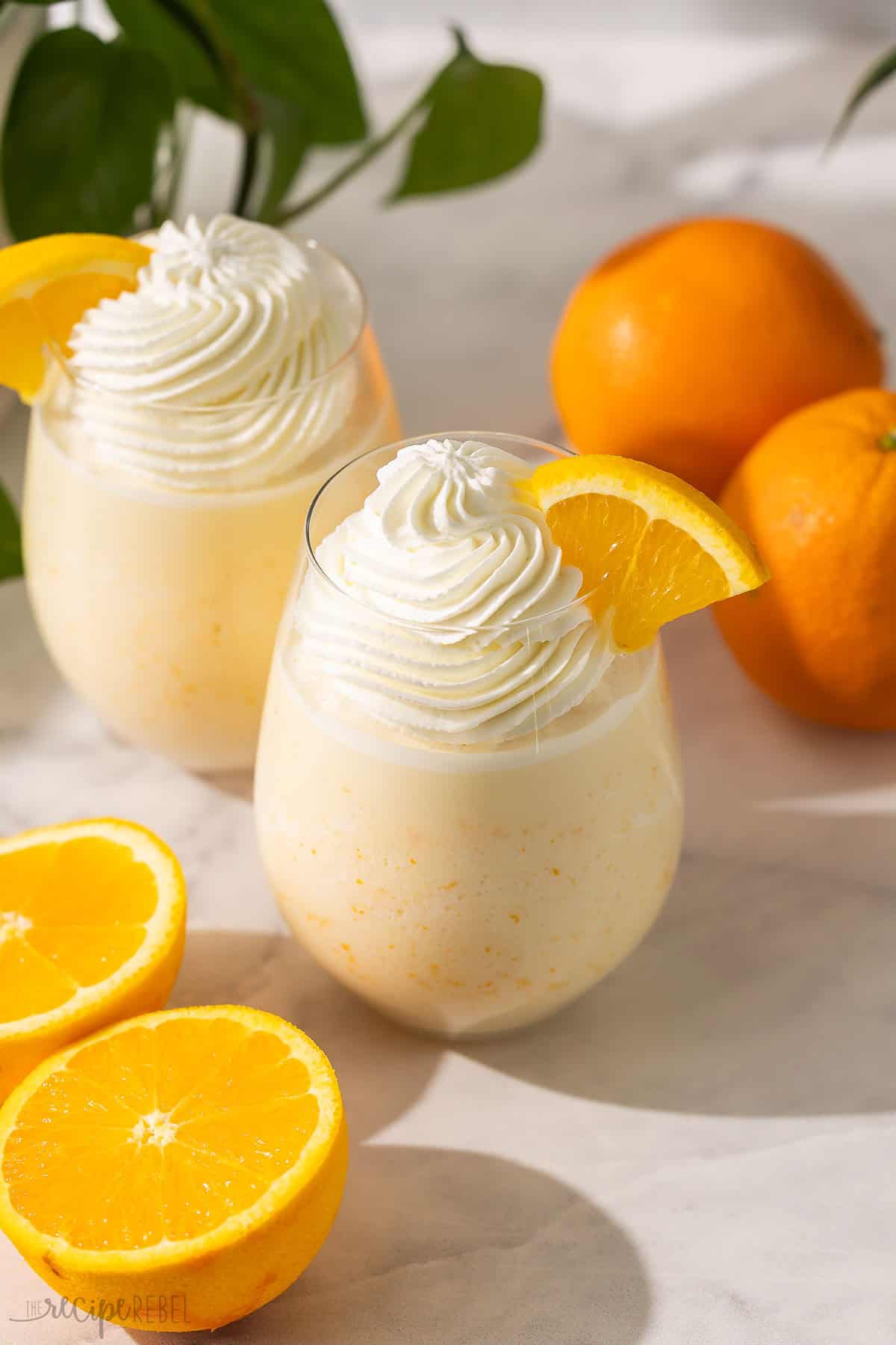 two glasses filled with orange creamsicle slushy topped whipped cream.