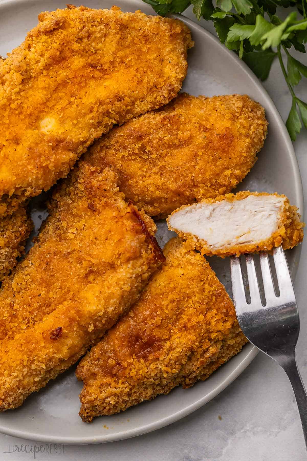 This easy Oven-Fried Chicken turns out perfectly crispy and juicy every time! The special seasoning blend tastes just like KFC, but it’s made with wholesome ingredients in your own kitchen. No deep fryer needed! #chicken #dinner #recipe | baked fried chicken | fried chicken breast | KFC copycat | chicken breast recipes | easy dinner ideas a piece of oven fried chicken on a fork with chicken beside it on a plate.