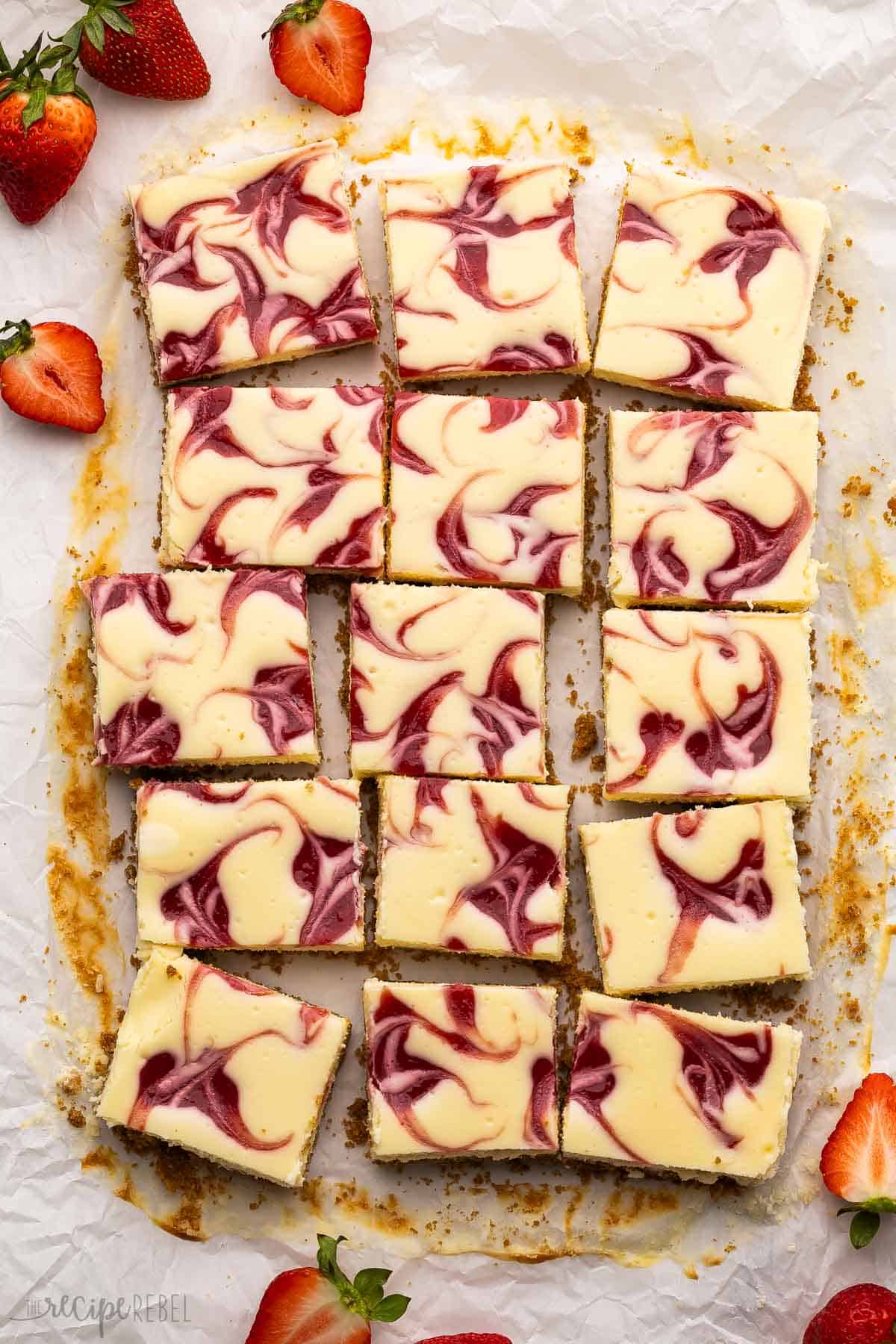 cut up strawberry swirl cheesecake bars on parchment paper surrounded by strawberries.