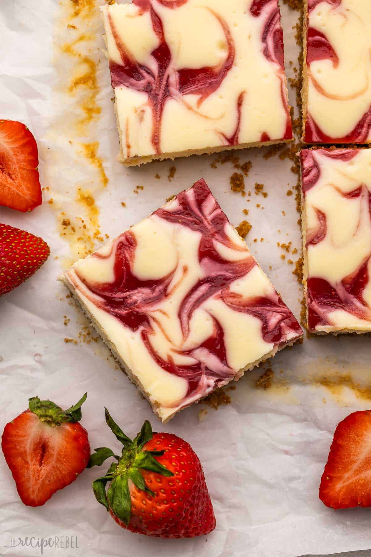 These Strawberry Cheesecake Bars are made with homemade strawberry filling, a tangy cheesecake base, and a buttery graham cracker crust. Made with fresh or frozen strawberries for a year-round treat! #strawberry #dessert #recipe | cheesecake recipe | baked cheesecake | dessert recipes | strawberry dessert | strawberry recipes cut up strawberry swirl cheesecake bars on parchment paper.