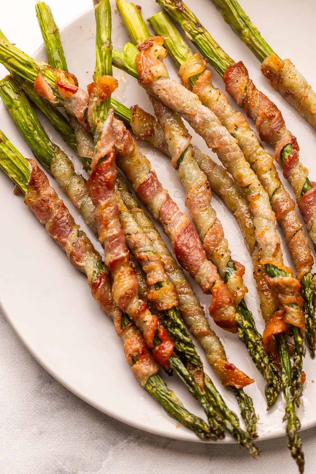 Bacon Wrapped Asparagus is an elegant yet easy side dish or appetizer made with slices of thin-cut bacon wrapped around asparagus stalks, sprinkled with seasonings, and baked until crispy. #appetizer #recipe | appetizer recipes | side dishes | vegetables | roast vegetables a white plate filled with bacon wrapped asparagus.