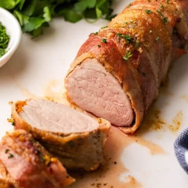 a partially sliced pork tenderloin on a white cutting board with parsley behind it.