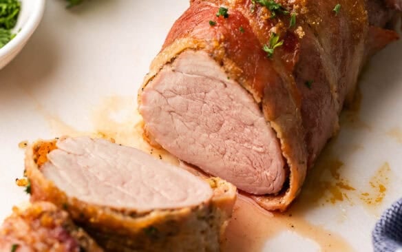 a partially sliced pork tenderloin on a white cutting board with parsley behind it.