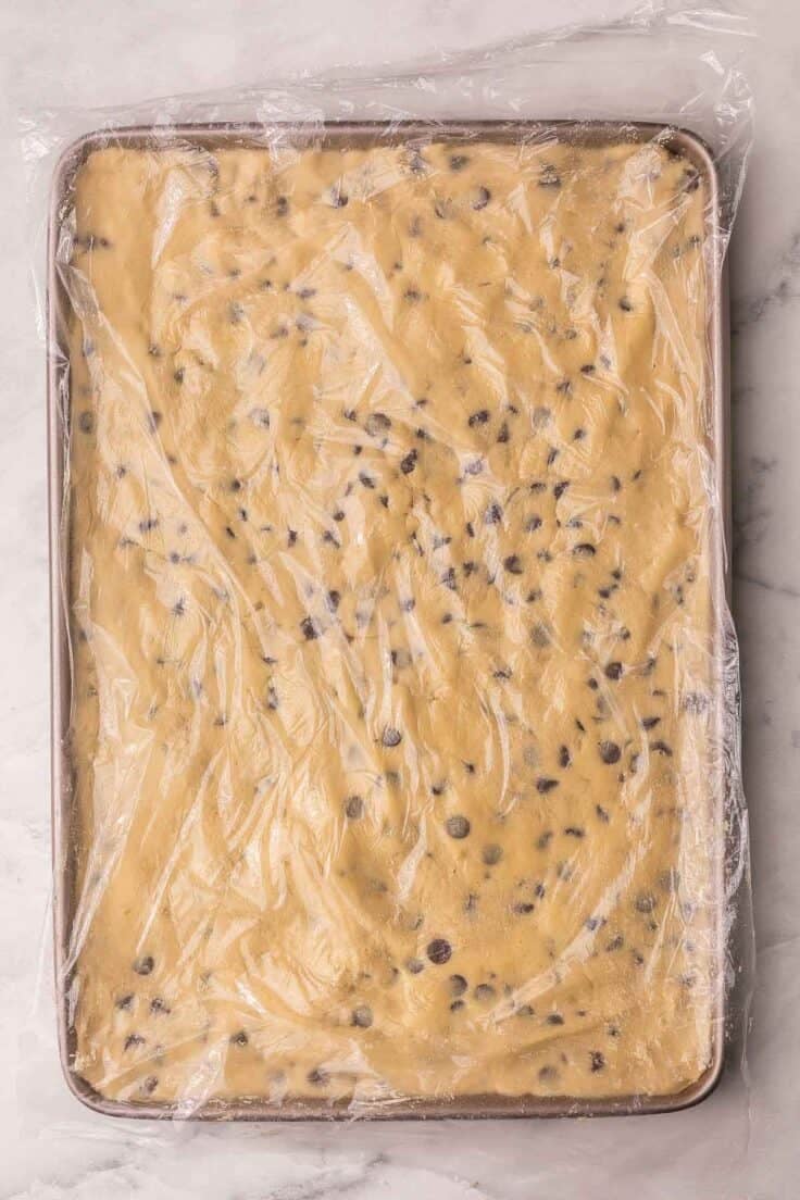large pan with chocolate chip cookie bar dough spread and covered with plastic wrap.