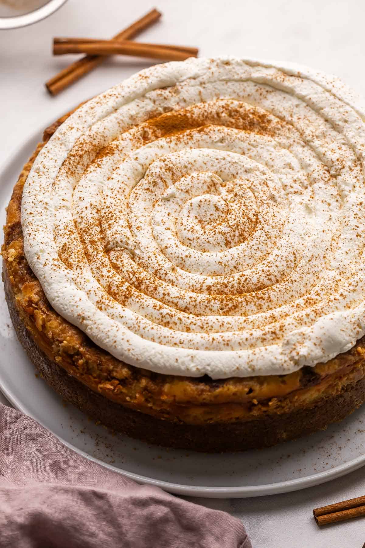 a full cinnamon roll cheesecake on a white plate with cinnamon sticks behind it.