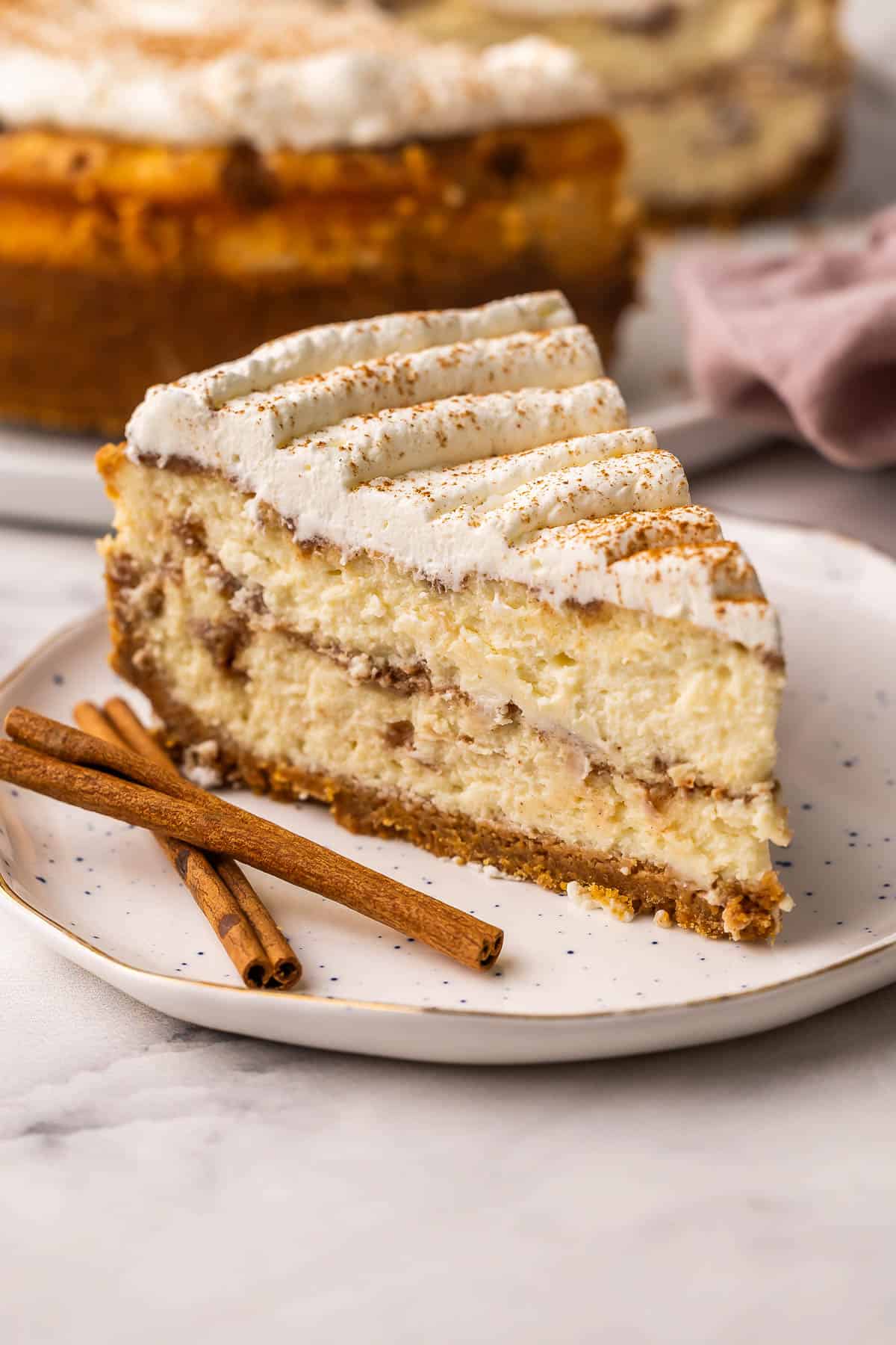 a slice of cinnamon roll cheesecake on a speckled plate with cinnamon sticks beside.