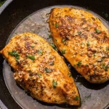 two pan fried chicken breasts in black skillet with garlic butter.