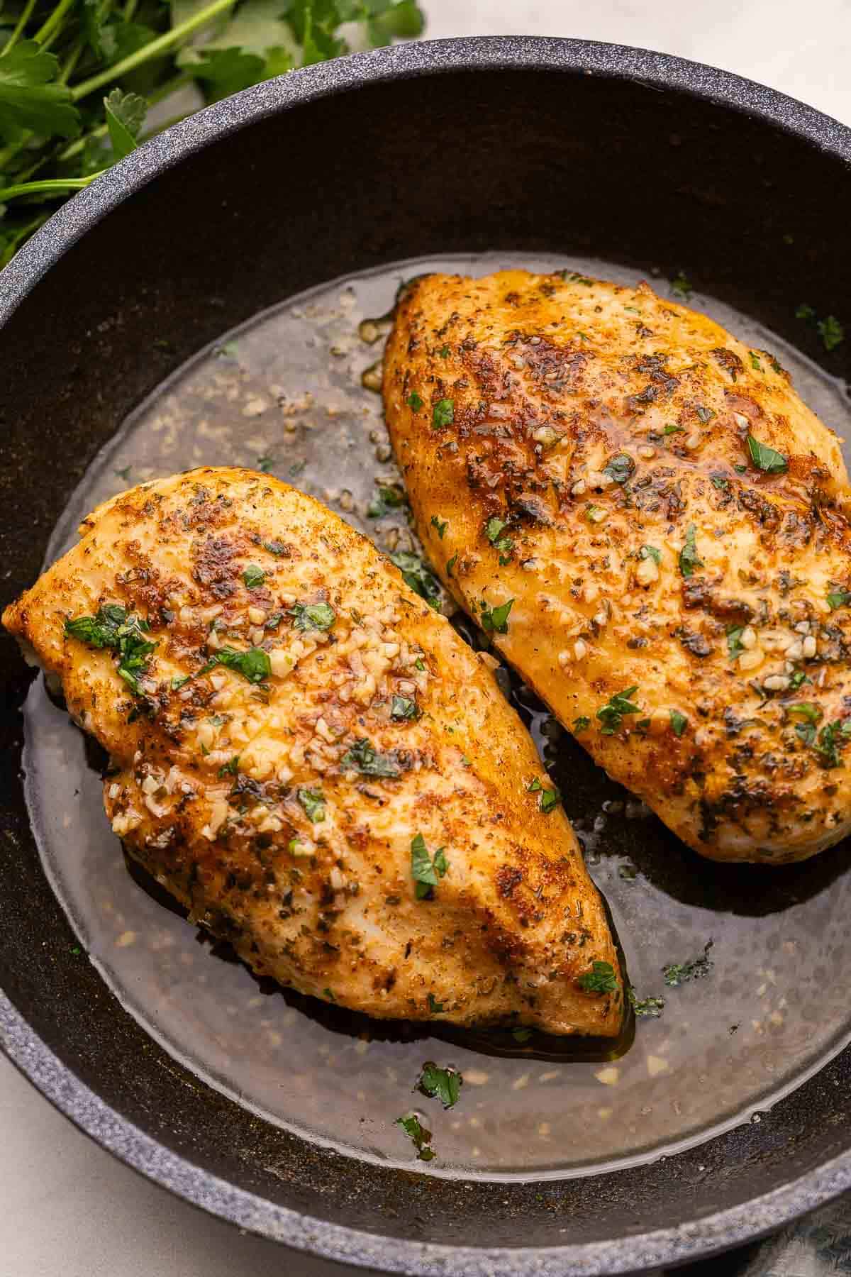 two pan fried chicken breasts in black skillet with garlic butter.