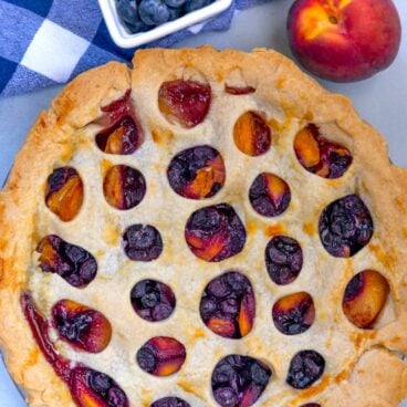 peach blueberry pie on a blue background with peaches and blueberries to the side.