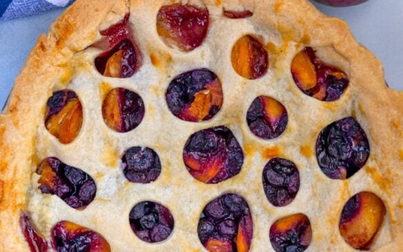 peach blueberry pie on a blue background with peaches and blueberries to the side.