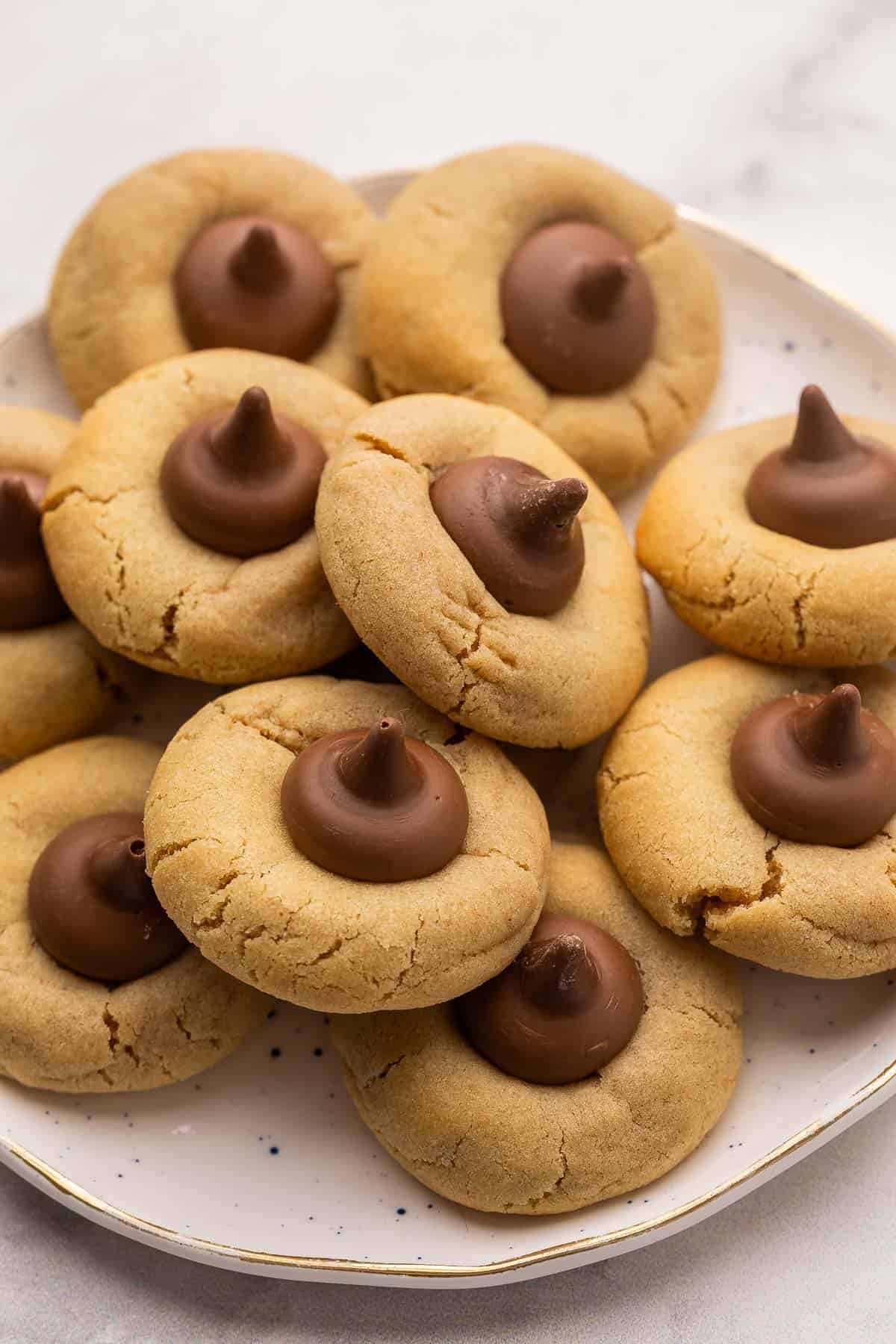 Peanut Butter Blossoms are made with a chewy peanut butter cookie base and a chocolate kiss pressed into the middle. They’re ready in just over 30 minutes and are the perfect nostalgic cookies for holidays, parties, or just because. #cookies #baking #dessert | cookie recipe | cookies | peanut butter cookies | peanut butter recipes | peanut butter chocolate a speckled plate with a bunch of peanut butter blossoms on it.