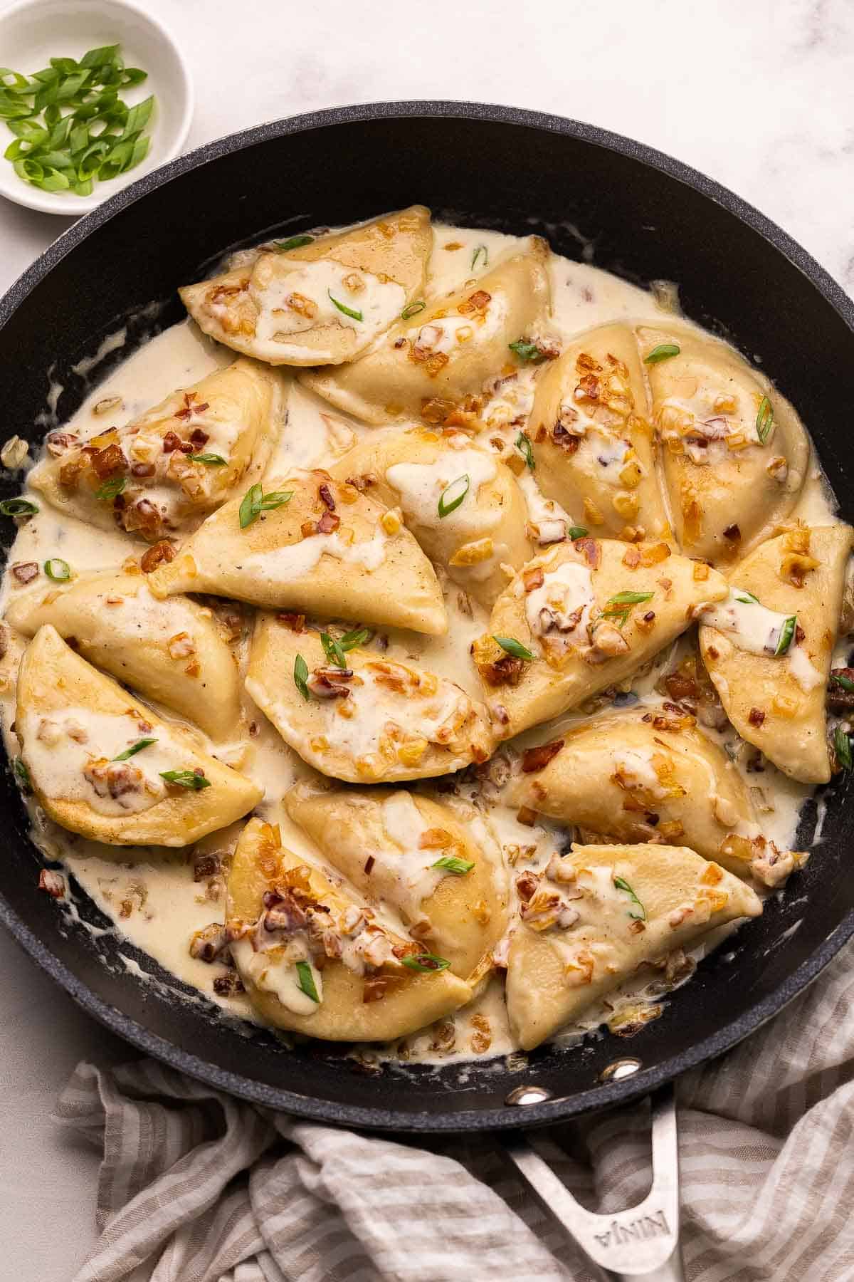 This Perogies recipe with Bacon and Onions recipe is made with frozen perogies swimming in a creamy sauce with smoky bacon and onions. Ready in 35 minutes, it's the perfect side dish for the holidays or whenever you're craving something comforting. | dinner | recipe | easy dinner ideas | pierogi | comfort food | skillet meals perogies with bacon and onions on large pan topped with chopped green onions.