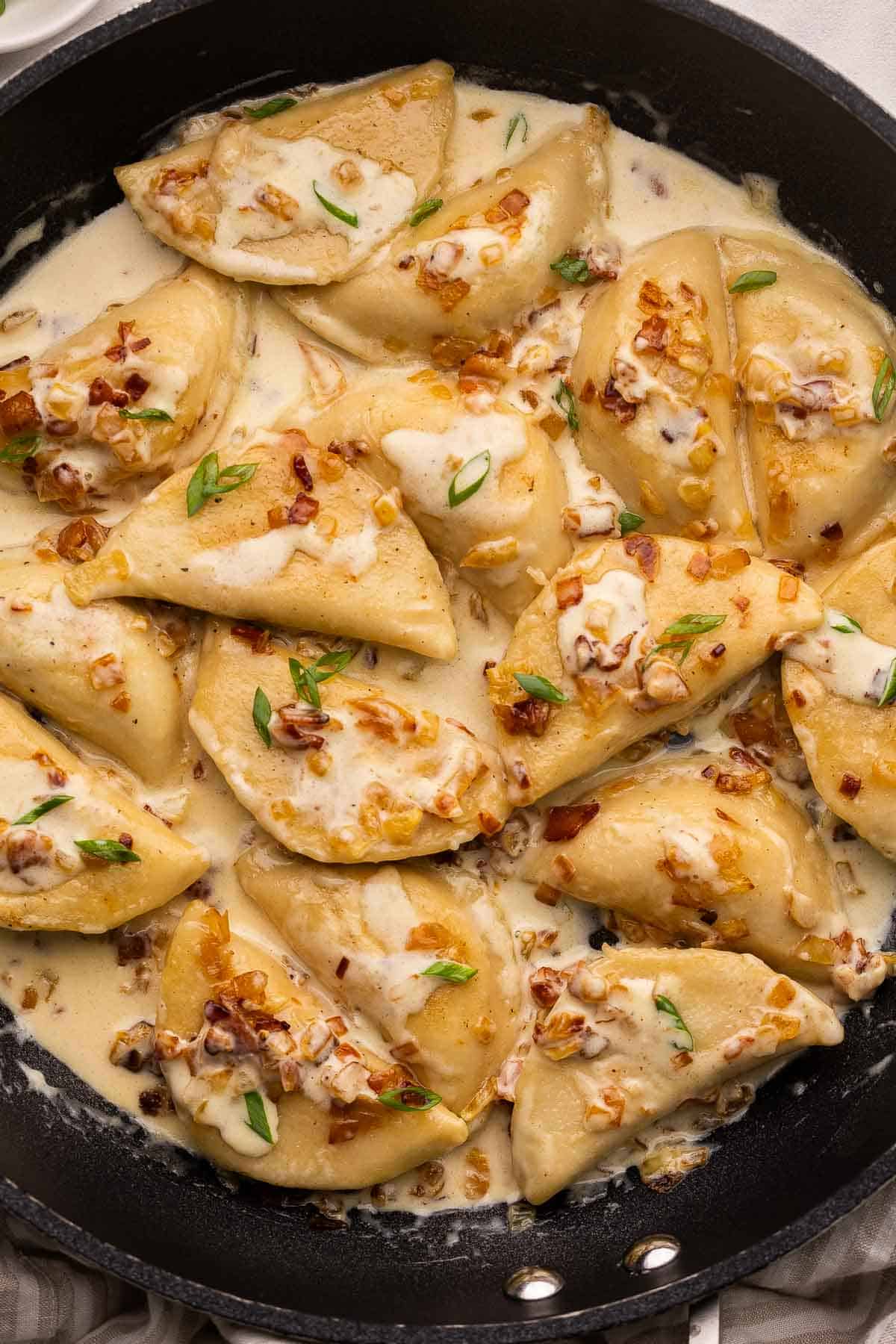 This Perogies recipe with Bacon and Onions recipe is made with frozen perogies swimming in a creamy sauce with smoky bacon and onions. Ready in 35 minutes, it's the perfect side dish for the holidays or whenever you're craving something comforting. | dinner | recipe | easy dinner ideas | pierogi | comfort food | skillet meals perogies covered in a creamy sauce in a large black pan.