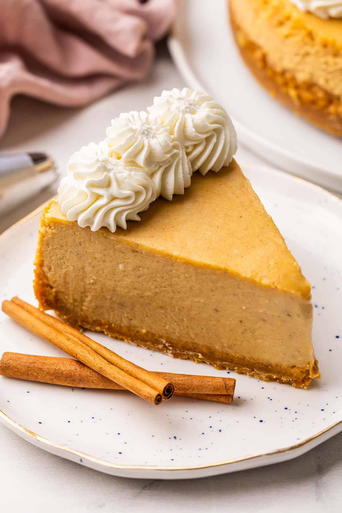 This baked Pumpkin Cheesecake has a super creamy filling and is packed with warm spices and cozy pumpkin flavor! It's easier than the rest, thanks to one secret method. No water bath and no cracks! #pumpkin #cheesecake #dessert | dessert recipes | pumpkin recipes | pumpkin dessert | baked cheesecake recipe | fall dessert | thanksgiving dessert a slice of pumpkin cheesecake on a speckled plate with cinnamon sticks beside.