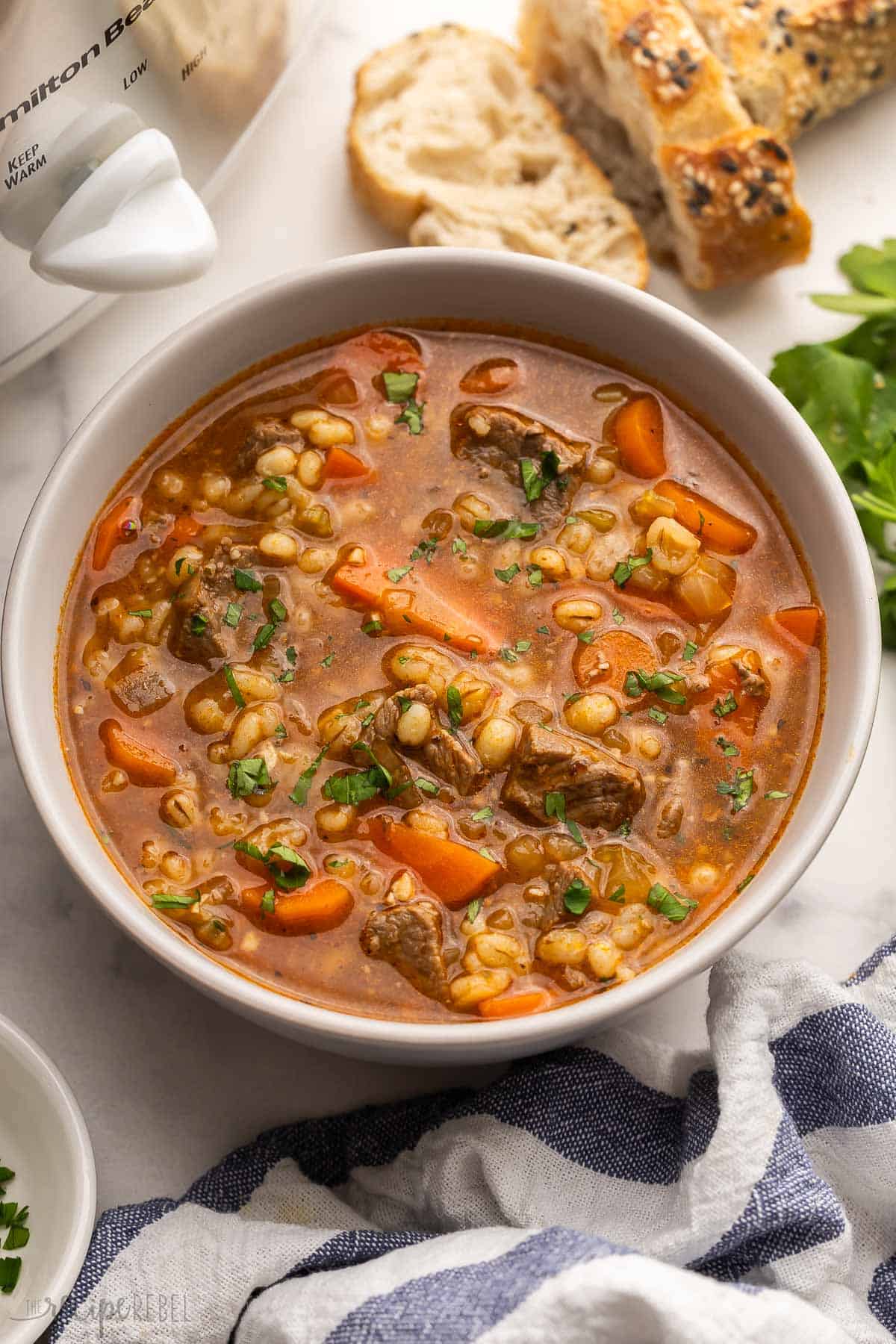 large bowl full of beef barley soup with bread in the background.