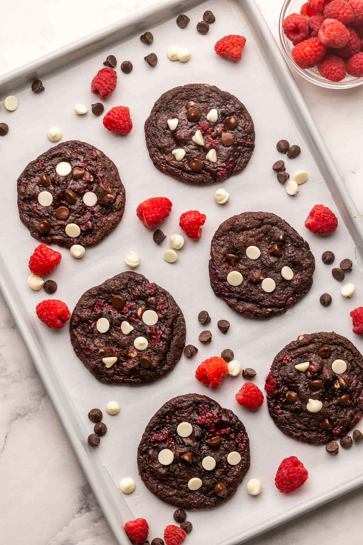 These Triple Chocolate Raspberry Cookies are made with double chocolate cookie dough, white chocolate chips, and tart raspberries! They're chewy, fudgy, and a little fruity, perfect for when you feel like changing things up. #chocolate #cookie #baking | baking recipes | chocolate cookie | chocolate cookies | double chocolate | baking from scratch | chocolate chip cookies a pan full of triple chocolate raspberry cookies with chocolate chips and raspberries beside them.