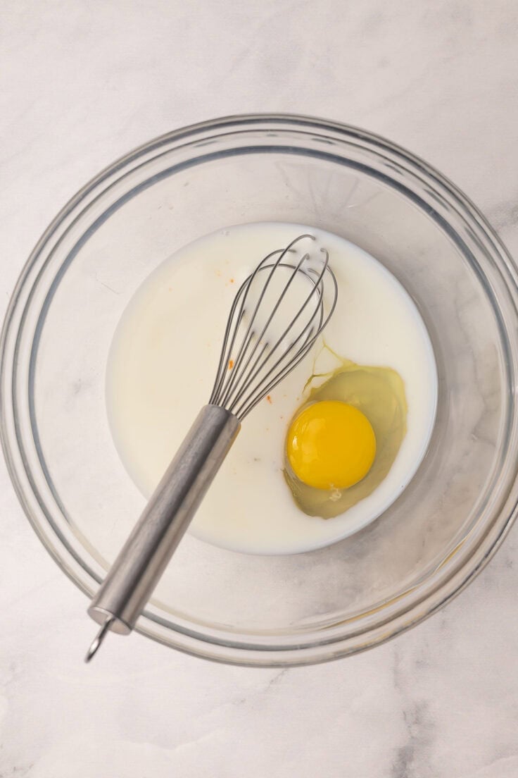 milk and an egg in a glass bowl with whisk.