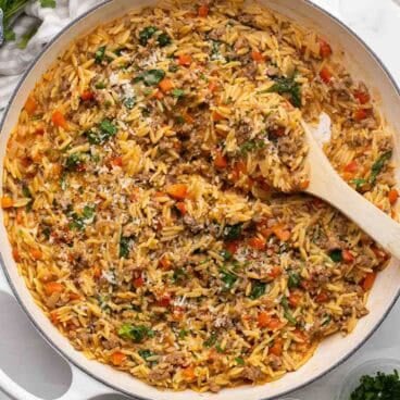 square image of italian sausage orzo in white braiser with wooden spoon stuck in.