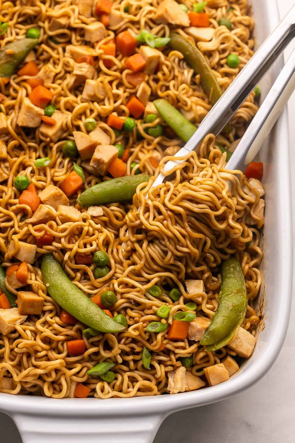 This Teriyaki Chicken Ramen Bake is a quick and easy 30-minute dinner with ramen noodles, tender cooked chicken breast, frozen veggies, and a sweet-savory sauce baked in one casserole dish! It's kid-friendly, customizable, and perfect for busy weeknights. #chicken #dinner | easy dinner ideas | chicken breast recipes | quick meals | easy recipes tongs lifting noodles out of white dish.