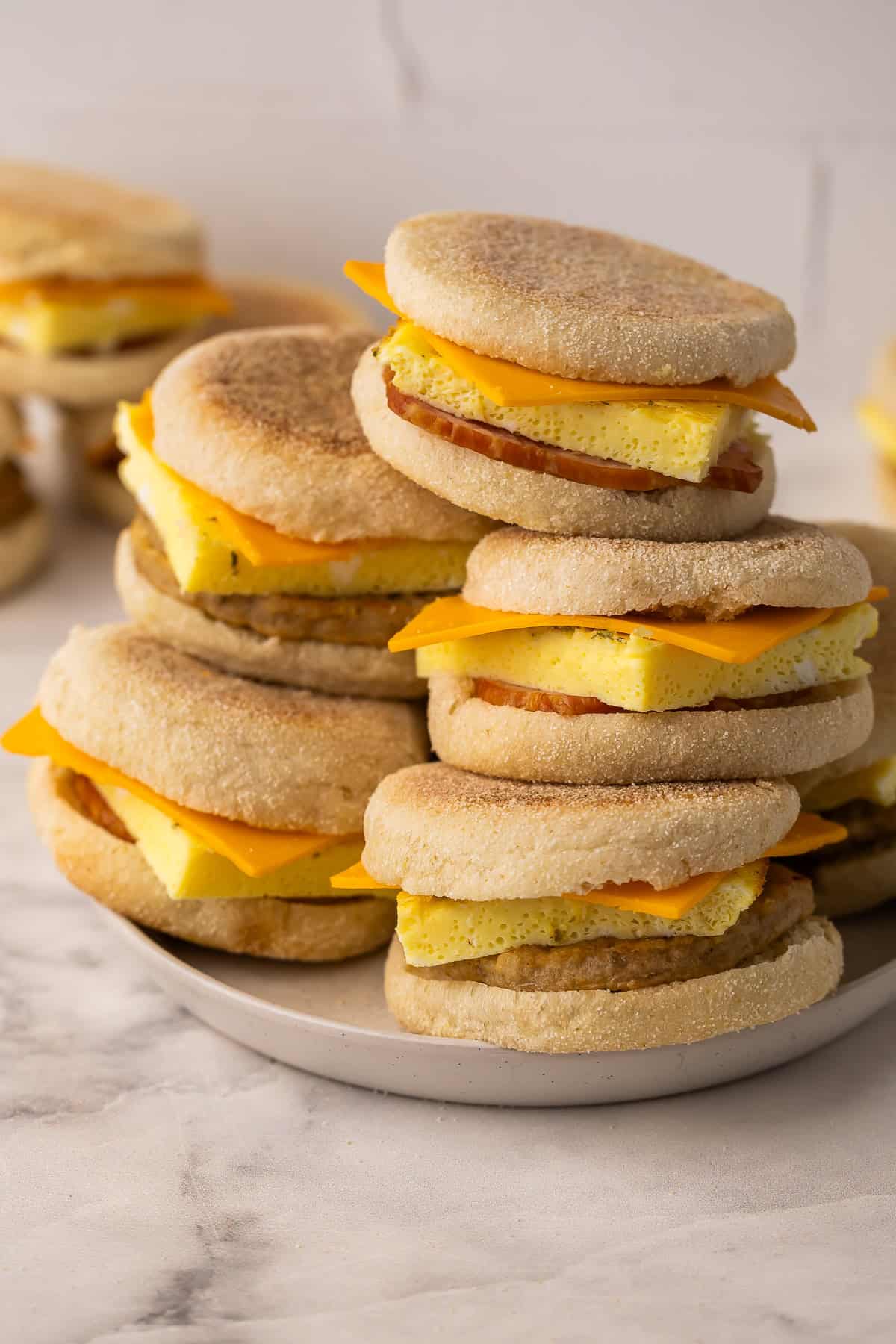 These Make Ahead Freezer Breakfast Sandwiches are hearty, cheesy, and freezer-friendly. Made with egg, cheese and your choice of meat, they're perfect for busy mornings, meal prep, or road trips! #freezermeal #breakfast #recipe | breakfast sandwich | freezer meals | breakfast prep | eggs | sausage | bacon | high protein a plate stacked high with breakfast sandwiches.