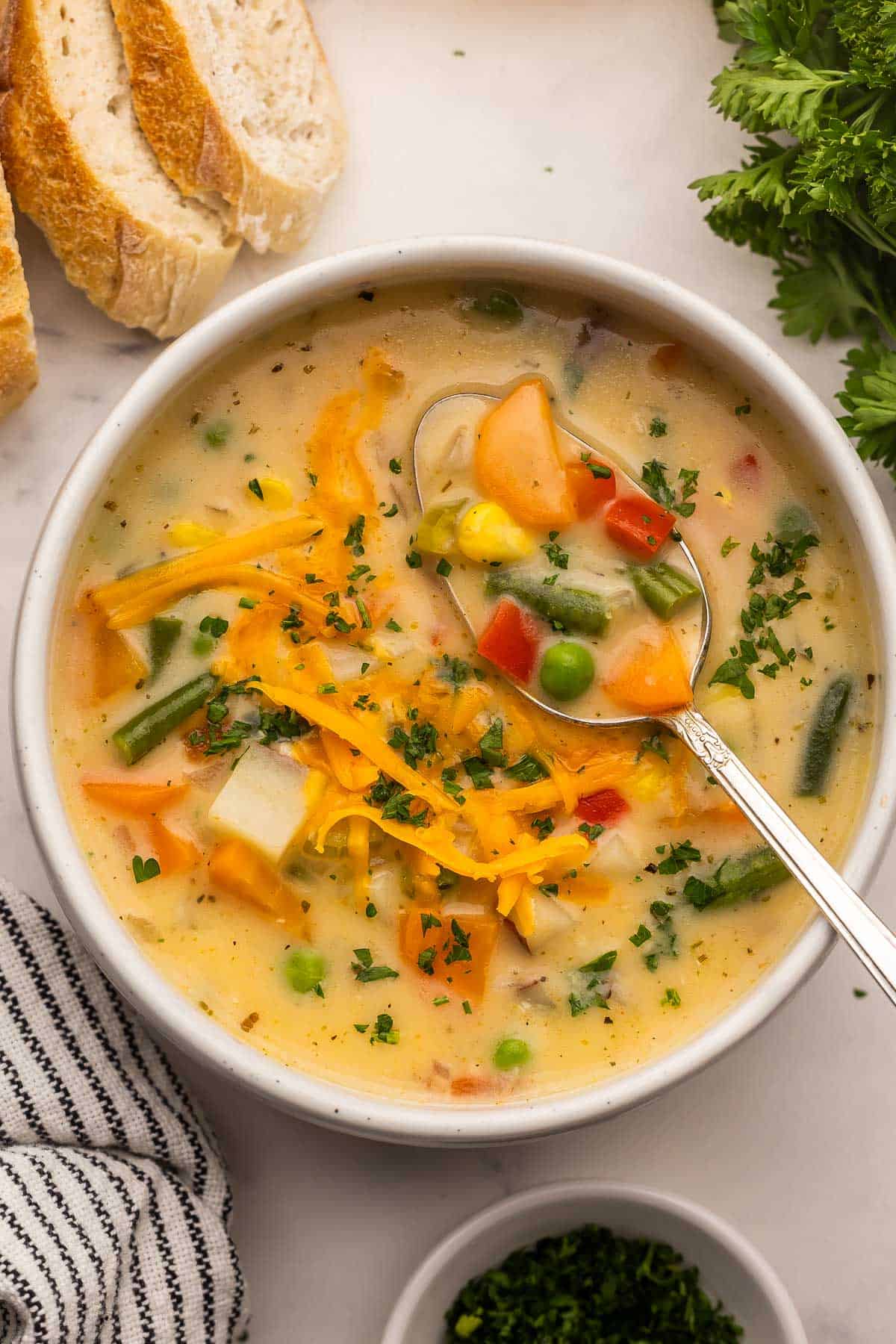 Cozy up with this thick and Creamy Vegetable Soup! Packed with potatoes, carrots, corn, peas, and melty cheddar, it’s a hearty 30-minute dinner the whole family loves (and our favorite way to eat our vegetables!). #soup #dinner #recipe | easy dinner ideas | easy dinner recipes | soup recipes | healthy soup | comfort food | vegetable soup | vegetable chowder | cheesy soup | creamy soups | healthy recipes a large bowl of vegetable soup topped with shredded cheese and chopped parsley.