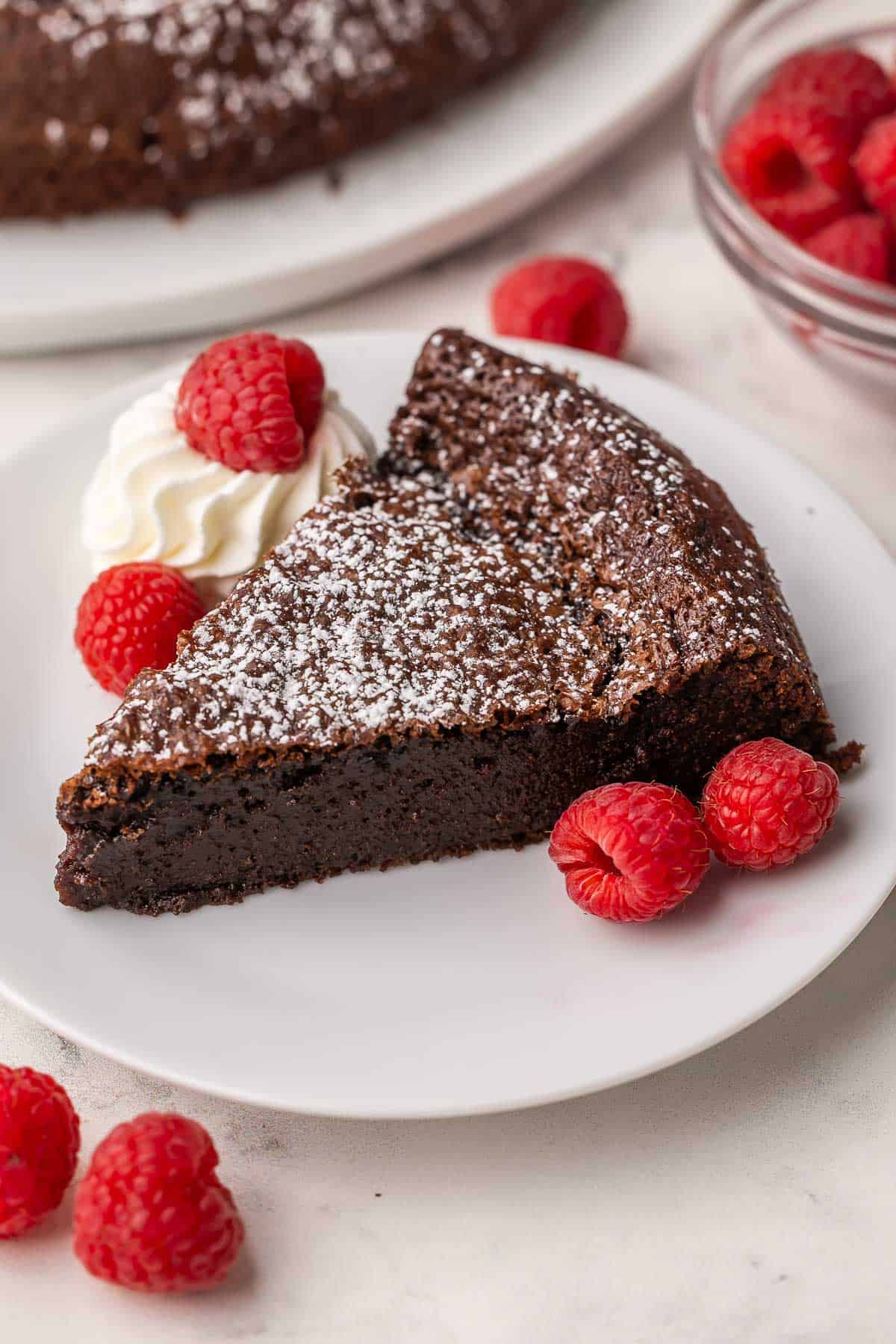 This Flourless Chocolate Cake is rich, fudgy, and naturally gluten-free. Easy to make with simple ingredients like melted chocolate, eggs and butter, it’s the perfect chocolate lover’s dessert! #chocolate #dessert #recipe | chocolate dessert | dessert recipes | the recipe rebel | easy recipes | valentine's day dessert | valentine's day recipe a slice of flourless chocolate cake on a white plate with whipped cream and raspberries.