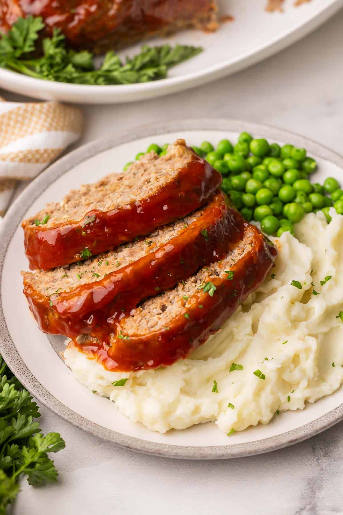 This truly is the BEST Meatloaf Recipe! It's juicy, flavorful, and topped with a sweet ketchup glaze. It's easy, comforting, and also makes a great freezer meal. #groundbeef #dinner #recipe | dinner ideas | dinner recipes | ground beef recipes | ground pork | comfort food | the recipe rebel | make ahead meals | freezer meals a plate with meatloaf, mashed potatoes and peas.